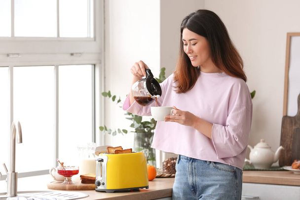 Beautiful young Asian woman drinking coffee and making tasty toasts in kitchen - Foto, Bild