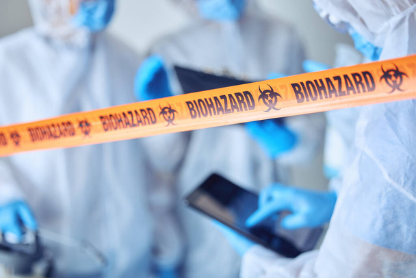 Closeup on biohazard quarantine tape. Hands of scientist using a digital tablet behind covid barrier. Medical professional working in a biohazard crime scene. Doctors working behind caution tape. - Zdjęcie, obraz