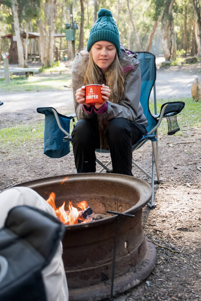 Camping lifestyle. Teenage girl wearing beanie hat sitting in a camping chair near campfire holding a red enamel mug. Happy camper - 写真・画像