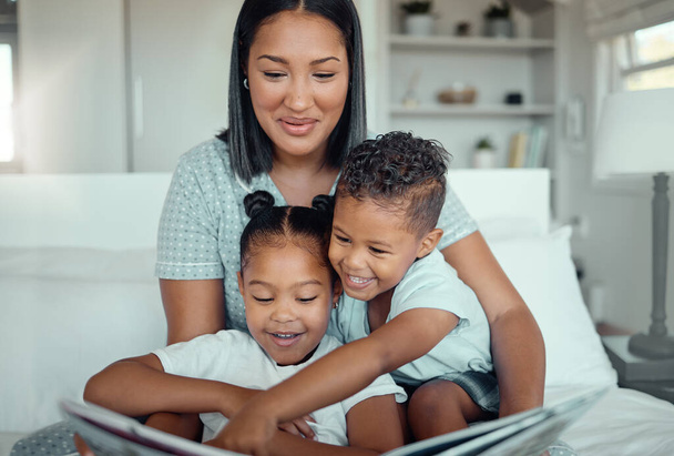 Young mixed race mother reading a storybook while relaxing at home with her two children. Smiling parent telling small kids funny fairy tale story kids pointing at book while they sit together. - Foto, imagen