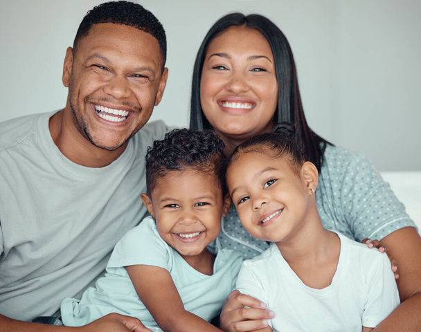 Portrait of a happy young mixed race family with two children wearing pyjamas and sitting at home together. Loving little sister and brother spending time with their parents on the weekend. - 写真・画像