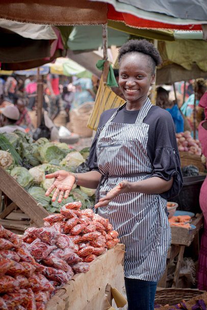 Una donna africana commerciante nigeriana, venditore, donna d'affari o imprenditore con un grembiule, mostrando felicemente i suoi beni esposti per attirare potenziali clienti in un mercato - Foto, immagini