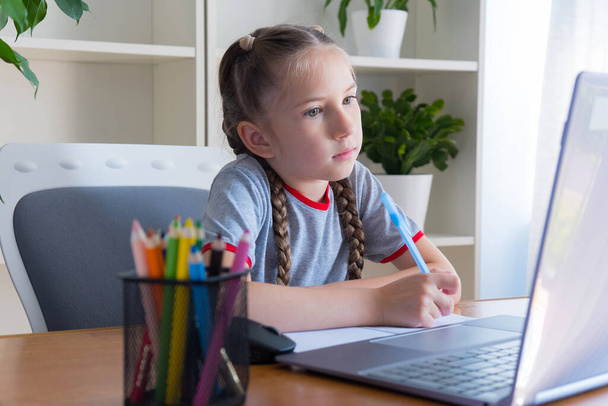 Egy 6-8 éves lány szürke pólóban, hogy otthon tanuljon a számítógépnél, tanuljon, házit csináljon, kommunikáljon az interneten, jegyzetfüzetbe írja. - Fotó, kép