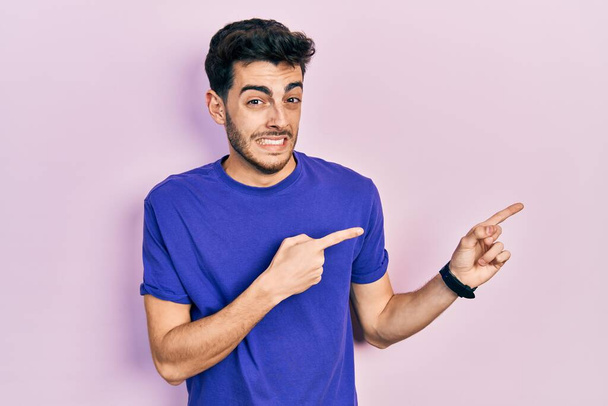 Jonge Spaanse man draagt casual t-shirt wijzen opzij bezorgd en nerveus met beide handen, bezorgd en verbaasd uitdrukking  - Foto, afbeelding