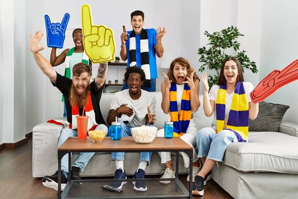 Group of friends supporting football team watching tv al home sitting on the sofa celebrating victory with happy smile and winner expression with raised hands  - Foto, imagen