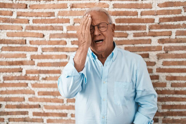 Senior man with grey hair standing over bricks wall yawning tired covering half face, eye and mouth with hand. face hurts in pain.  - Foto, afbeelding