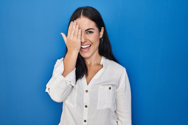 Молода іспаномовна жінка стоїть над синім тлом, покриваючи одне око рукою, впевнена посмішка на обличчі і несподівані емоції
.  - Фото, зображення