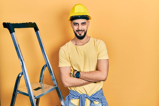 Handsome man with beard by construction stairs wearing hardhat happy face smiling with crossed arms looking at the camera. positive person.  - Foto, Imagem
