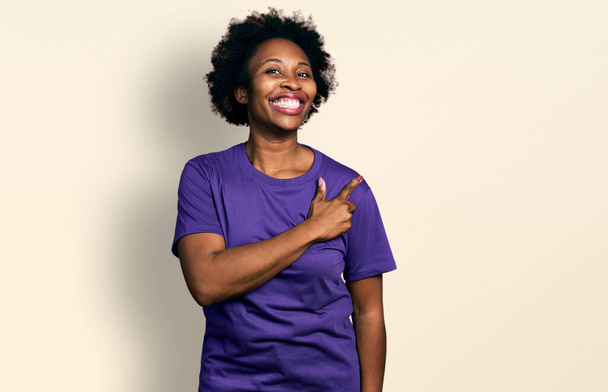 Afrikaans amerikaanse vrouw met afro haar dragen casual paars t-shirt vrolijk met een glimlach op het gezicht wijzen met de hand en vinger naar de zijkant met vrolijke en natuurlijke expressie  - Foto, afbeelding
