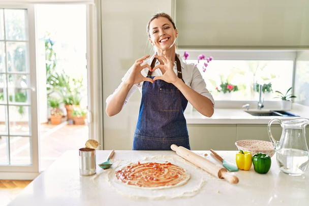 Красива блондинка в фартусі готує піцу, посміхаючись, показуючи серцевий символ і форму руками. романтична концепція
.  - Фото, зображення