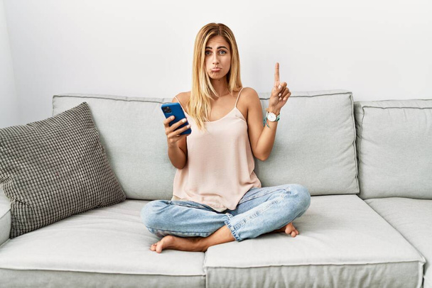 Blonde schöne junge Frau sitzt auf dem Sofa zu Hause mit Smartphone zeigt nach oben Blick traurig und aufgebracht, zeigt die Richtung mit den Fingern, unglücklich und deprimiert.  - Foto, Bild