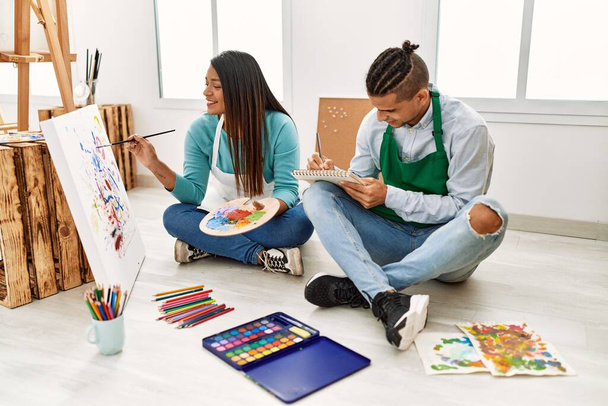 Młoda Latynoska para malarzy uśmiechająca się szczęśliwa siedząc na podłodze malując w pracowni artystycznej. - Zdjęcie, obraz