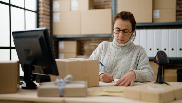 Middle age hispanic woman ecommerce business worker writing on book at storehouse office - Φωτογραφία, εικόνα