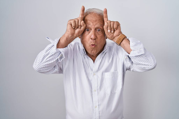 Starší muž s šedými vlasy stojící nad izolovaným pozadím dělá legrační gesto s prstem přes hlavu jako býčí rohy  - Fotografie, Obrázek
