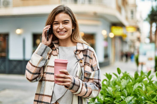 Молодая блондинка держит кофе разговаривая по смартфону на улице - Фото, изображение