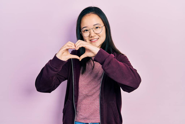 Молода китайська дівчинка у повсякденному одязі і окулярах посміхається в коханні, показуючи символ і форму серця руками. романтична концепція.  - Фото, зображення