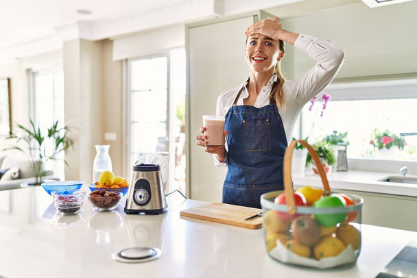 Beautiful blonde woman wearing apron drinking smoothie at the kitchen stressed and frustrated with hand on head, surprised and angry face  - Fotó, kép
