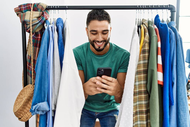 Νεαρός πελάτης arab man χρησιμοποιώντας smartphone που εμφανίζεται μέσω σχάρα ρούχων στο κατάστημα ρούχων - Φωτογραφία, εικόνα