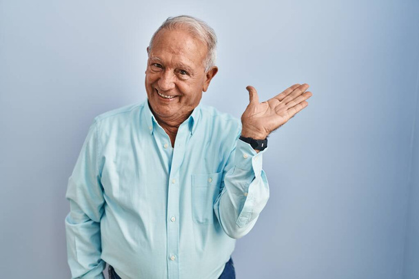 Senior man met grijs haar staan over blauwe achtergrond glimlachend vrolijke presenteren en wijzen met palm van de hand kijken naar de camera.  - Foto, afbeelding