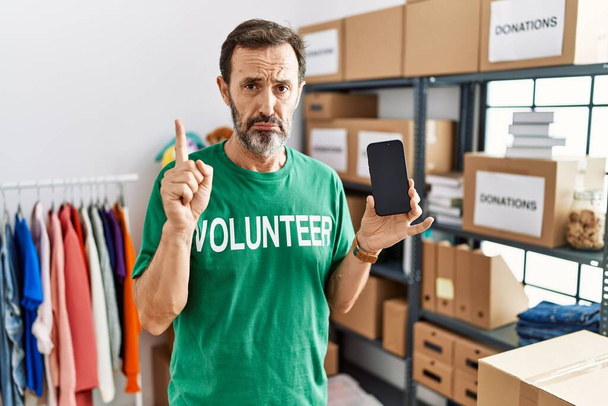 Middle age man with beard wearing volunteer t shirt holding smartphone pointing up looking sad and upset, indicating direction with fingers, unhappy and depressed.  - Photo, image