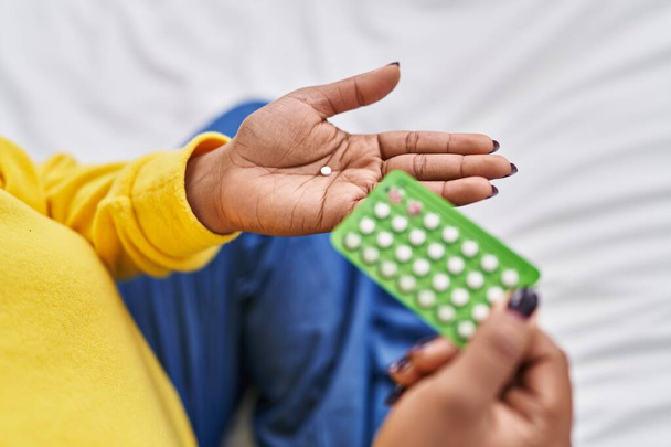Africano americano segurando pílulas anticoncepcionais sentado na cama no quarto - Foto, Imagem