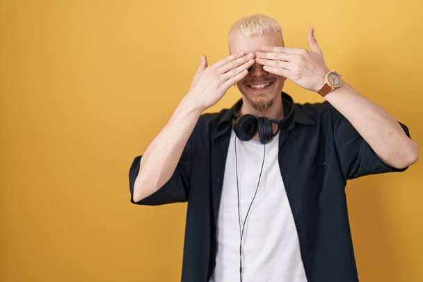 Young caucasian man wearing sunglasses standing over yellow background covering eyes with hands smiling cheerful and funny. blind concept.  - Foto, Imagen