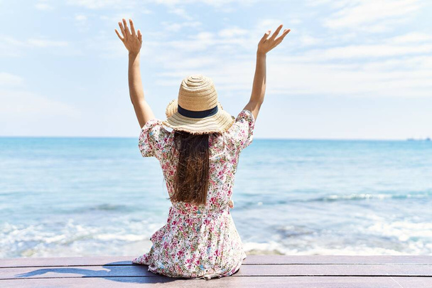 Νεαρή Λατίνα στην πίσω όψη φορώντας καλοκαιρινό καπέλο καθισμένη στον πάγκο στην παραλία. - Φωτογραφία, εικόνα