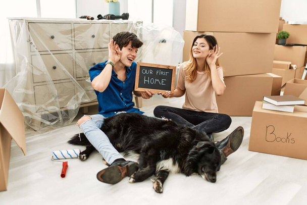 Young caucasian couple with dog holding our first home blackboard at new house smiling with hand over ear listening an hearing to rumor or gossip. deafness concept.  - Photo, image