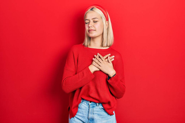 Beautiful blonde woman wearing casual red sweater smiling with hands on chest with closed eyes and grateful gesture on face. health concept.  - Photo, Image