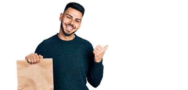 Young hispanic man with beard holding take away paper bag pointing thumb up to the side smiling happy with open mouth  - Photo, Image
