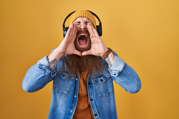 Caucasian man with long beard listening to music using headphones shouting angry out loud with hands over mouth  - Фото, изображение