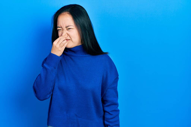 Young chinese girl wearing casual clothes smelling something stinky and disgusting, intolerable smell, holding breath with fingers on nose. bad smell  - Фото, зображення