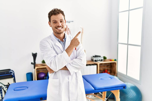 Handsome young man working at pain recovery clinic with a big smile on face, pointing with hand and finger to the side looking at the camera.  - Fotoğraf, Görsel
