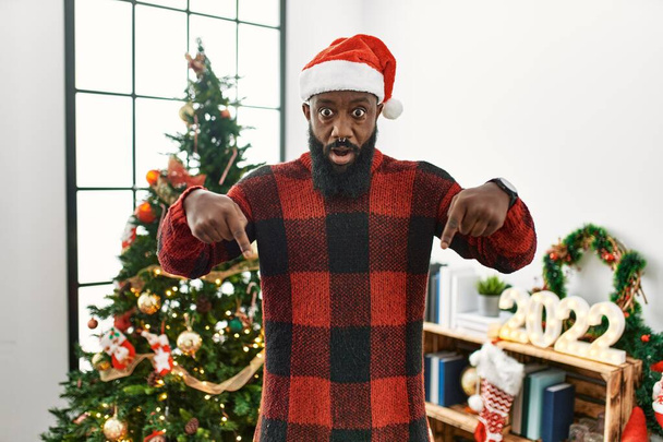 Αφροαμερικάνος που φοράει καπέλο Σάντα Κλάους και στέκεται δίπλα στο χριστουγεννιάτικο δέντρο δείχνοντας τα δάχτυλά του με διαφημίσεις, έκπληκτο πρόσωπο και ανοιχτό στόμα.  - Φωτογραφία, εικόνα
