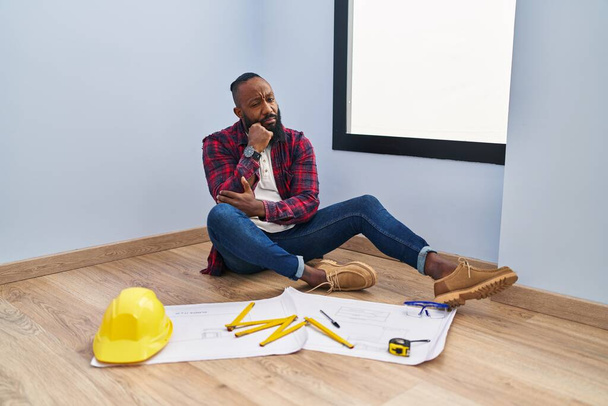 Αφροαμερικάνος που κάθεται στο πάτωμα στο νέο σπίτι κοιτάζοντας τα σχέδια με το χέρι στο πηγούνι σκεπτόμενος την ερώτηση, την σκεπτική έκφραση. Χαμογελώντας με ευγενικό πρόσωπο. έννοια της αμφιβολίας.  - Φωτογραφία, εικόνα