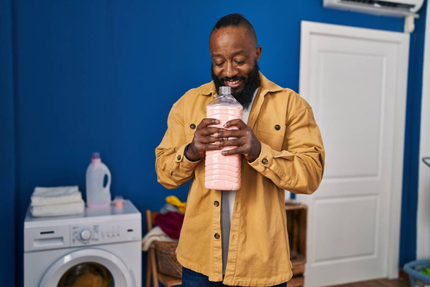 Young african american man smilng confident smelling detergent at laundry room - Zdjęcie, obraz