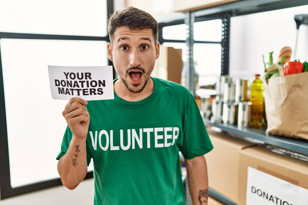 Young volunteer man holding your donation matters banner scared and amazed with open mouth for surprise, disbelief face  - Valokuva, kuva