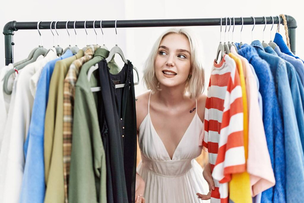 Jonge blanke vrouw glimlachend zelfverzekerd verschijnen door kleding in de kledingwinkel - Foto, afbeelding