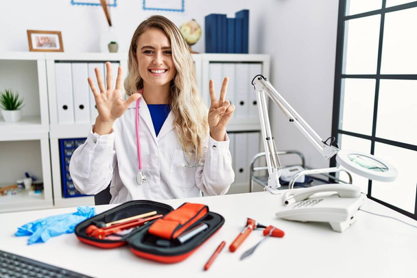 Fiatal gyönyörű orvos nő reflex kalapáccsal és orvosi műszerekkel mutatja és mutat fel a hetes számú ujjaival, miközben mosolyog magabiztos és boldog.  - Fotó, kép