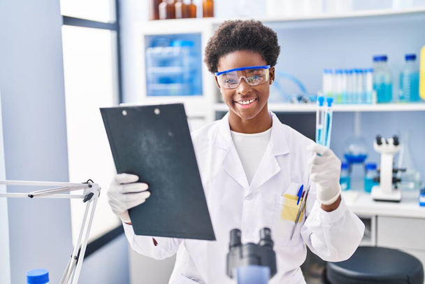 African american woman wearing scientist uniform reading document holding test tubes at laboratory - Foto, Bild