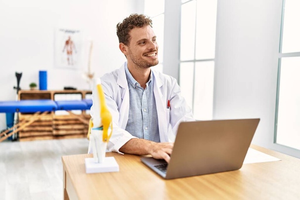 Fiatal spanyol férfi fizioterapeuta egyenruhát visel laptoppal dolgozik a klinikán. - Fotó, kép