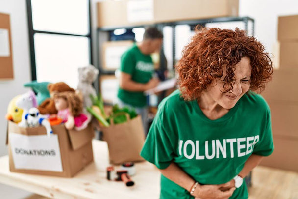 Middle age woman wearing volunteer t shirt at donations stand with hand on stomach because nausea, painful disease feeling unwell. ache concept.  - Foto, Imagen
