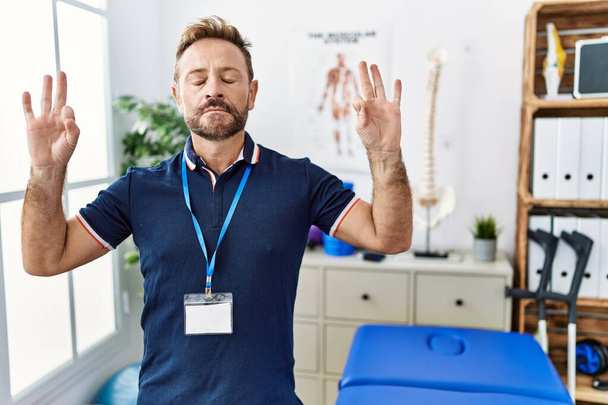 Middle age physiotherapist man working at pain recovery clinic relax and smiling with eyes closed doing meditation gesture with fingers. yoga concept.  - Photo, image