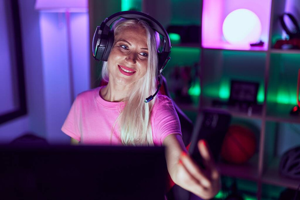 jong blond vrouw streamer dragen van hoofdtelefoon maken selfie door smartphone in gaming kamer - Foto, afbeelding