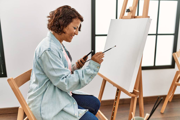 Middle age hispanic artist woman using smartphone drawing at art studio. - Photo, Image