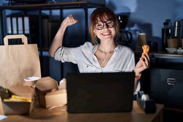 Young beautiful woman working using computer laptop and eating delivery food strong person showing arm muscle, confident and proud of power  - Фото, изображение