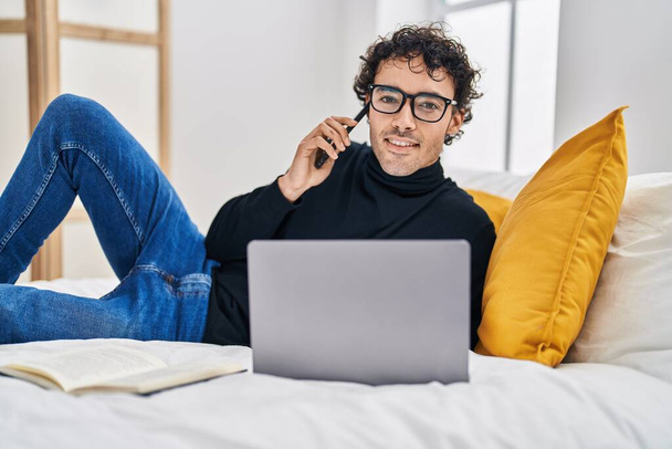Молодий іспаномовний чоловік використовує ноутбук, розмовляючи на смартфоні в спальні
 - Фото, зображення
