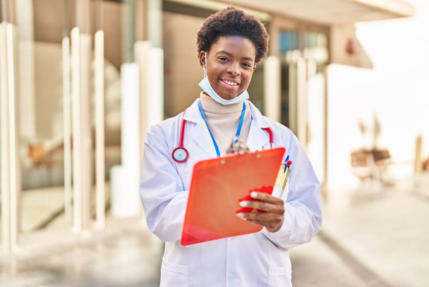 African american woman wearing doctor uniform writing on clipboard at street - Foto, afbeelding