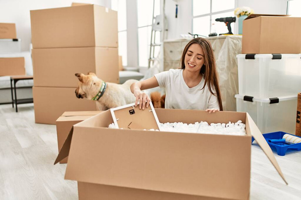 Young hispanic woman unboxing package sitting on floor with dog at new home - 写真・画像