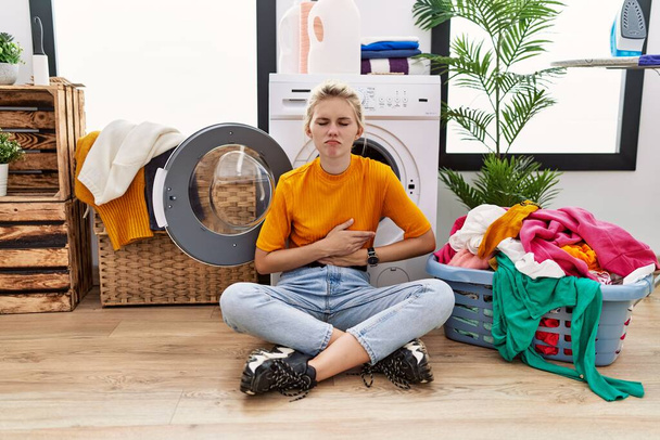 Young blonde woman doing laundry sitting by washing machine with hand on stomach because indigestion, painful illness feeling unwell. ache concept.  - Foto, Imagen
